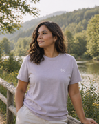 Woman wearing a light purple t-shirt with a logo, standing outdoors near a forest and lake.