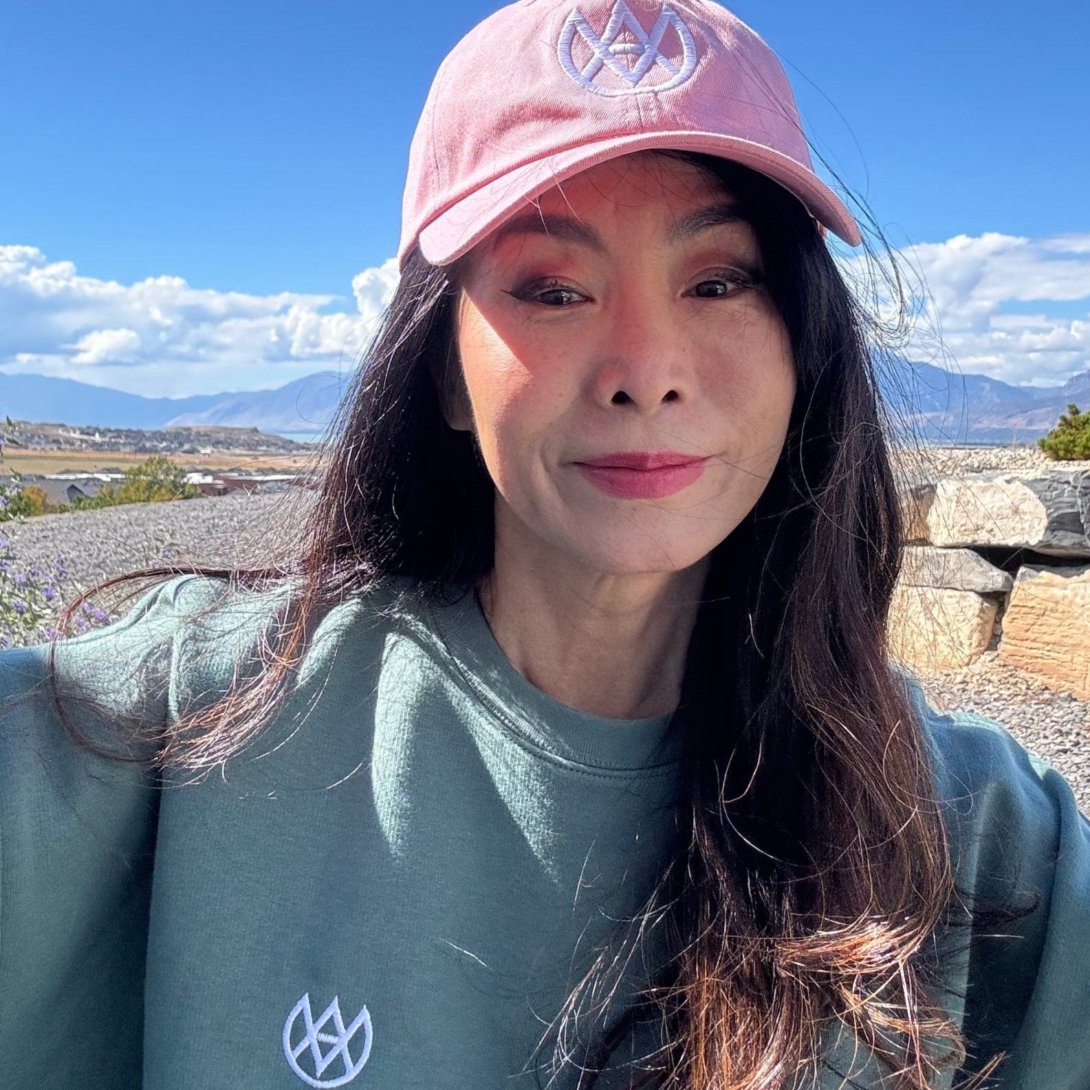 Person wearing a pink cap and green shirt with a logo, standing outdoors with mountains in the background.